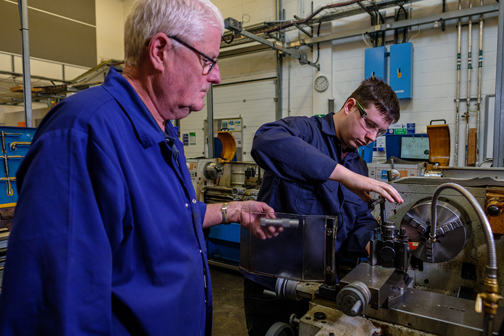 Older man passing a piece of equipment to a young man working at a machine in a workshop