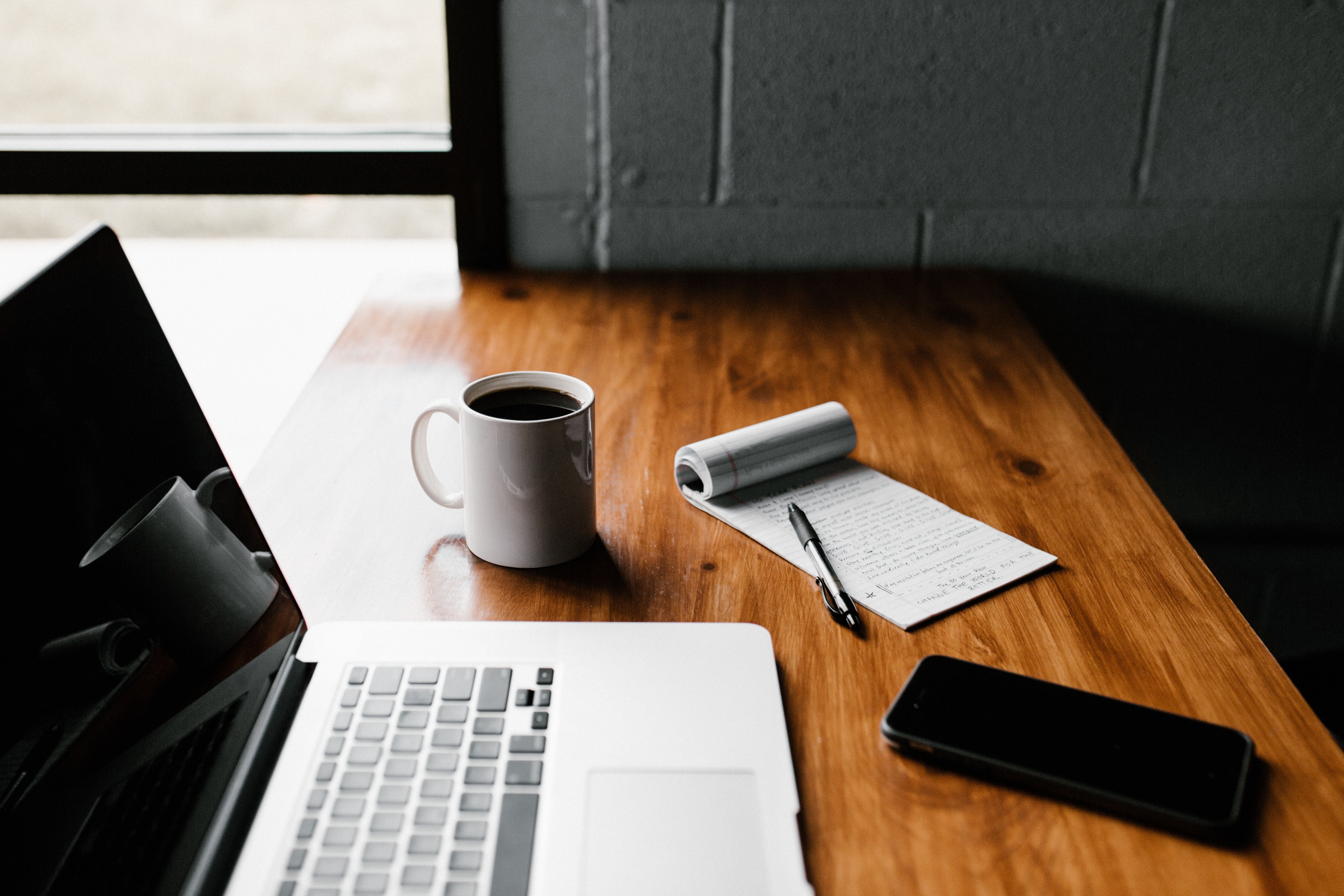 A laptop computer, mobile phone, note pad, pen and cup of coffee on a desk