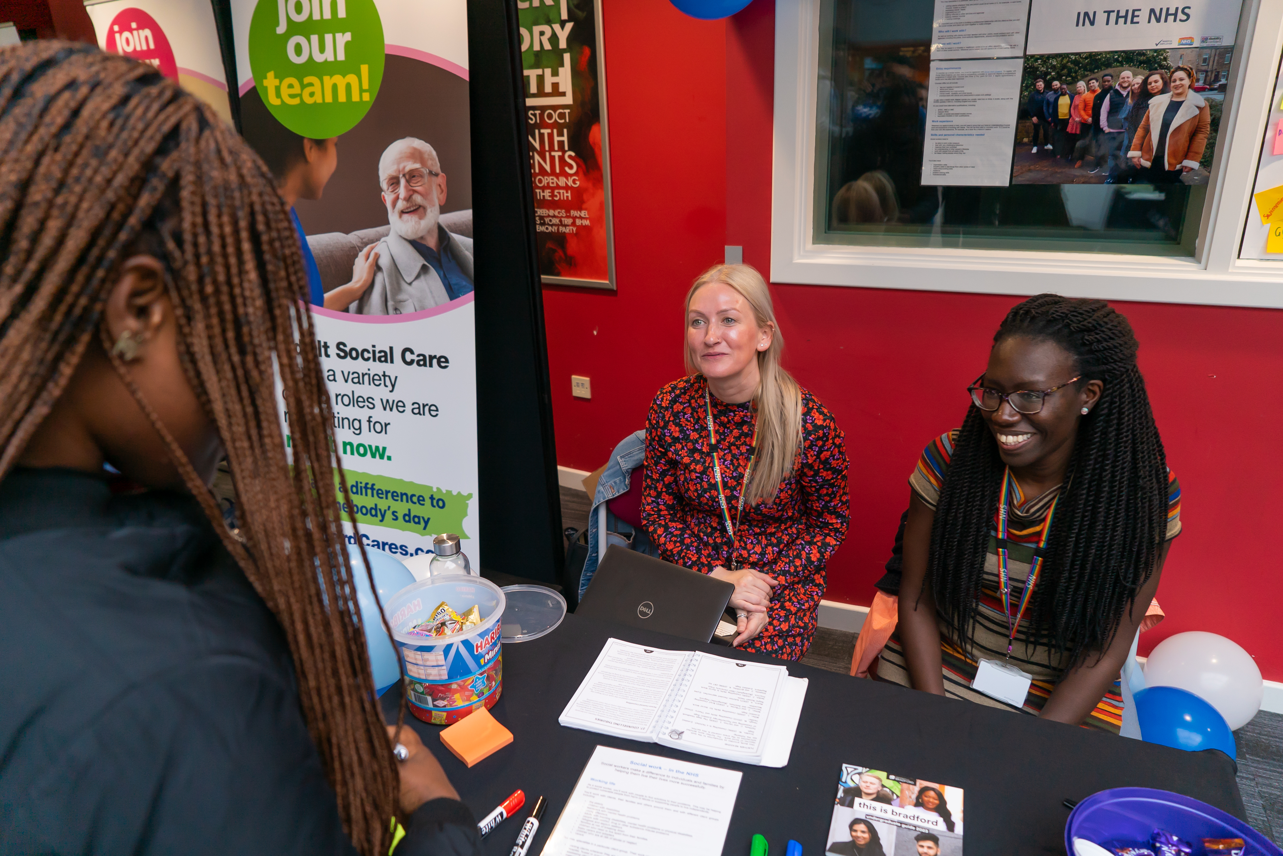 Two women sat at a desk talking to a woman standing, with a banner behind about social care
