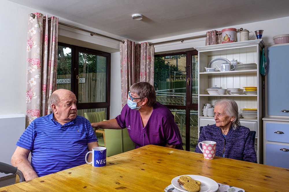 Two older people sit at a table talking to a younger woman in healthcare uniform