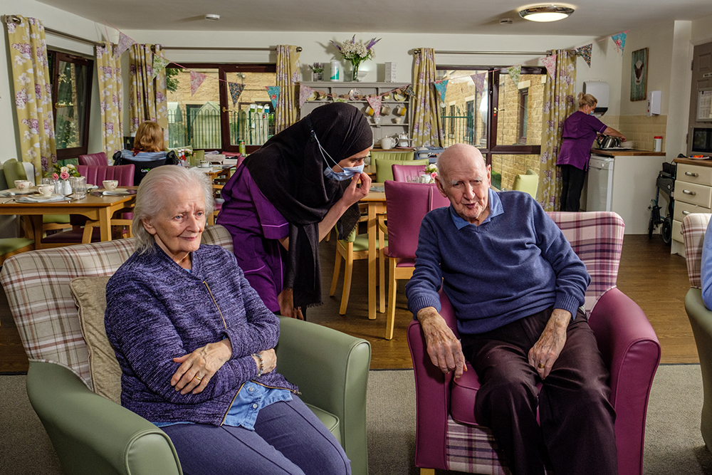 An older man and woman sat in armchairs talking to a woman in healthcare uniform
