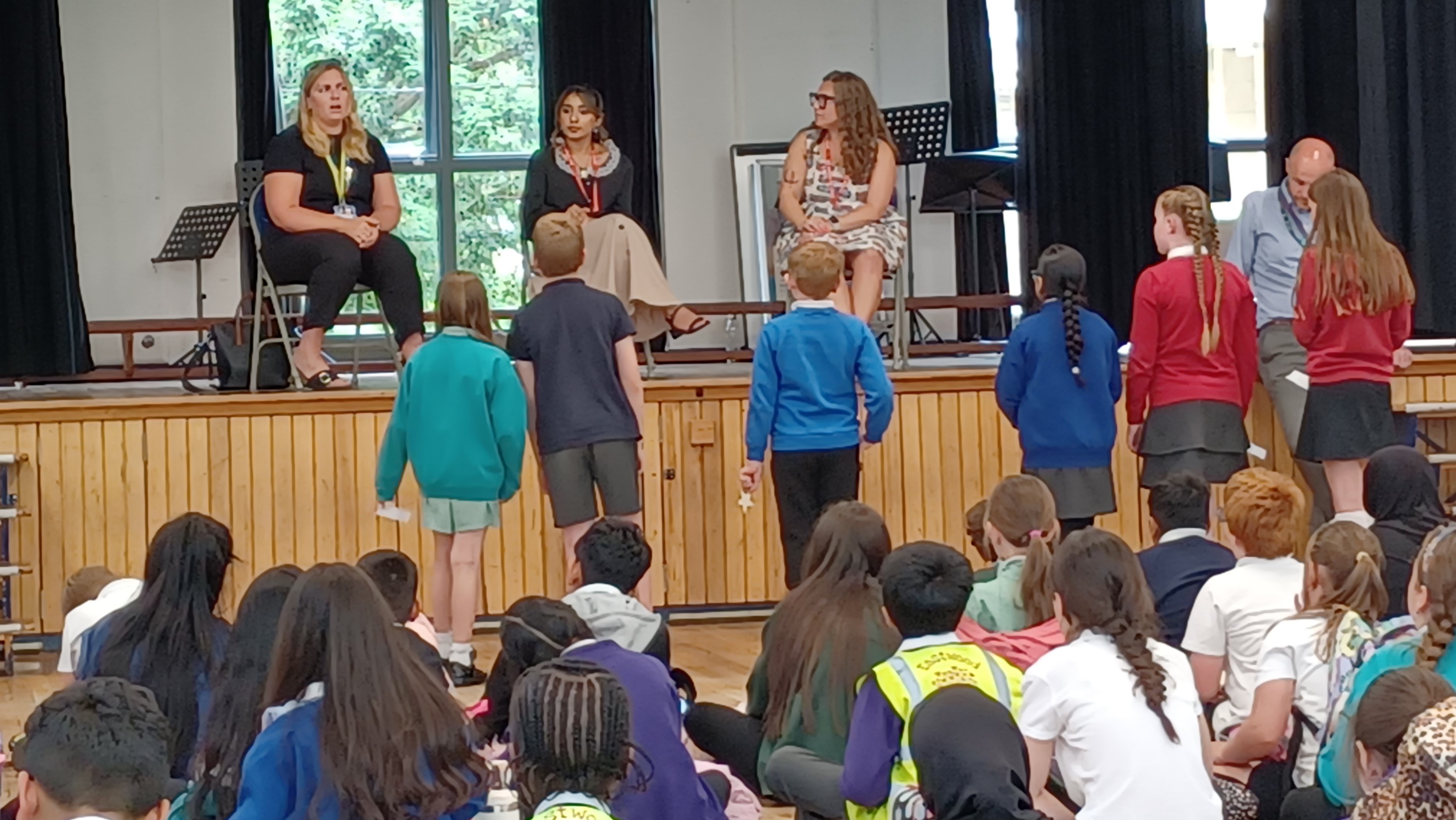 A large group of school children sat in a hall, some standing at the front, listening to three speakers