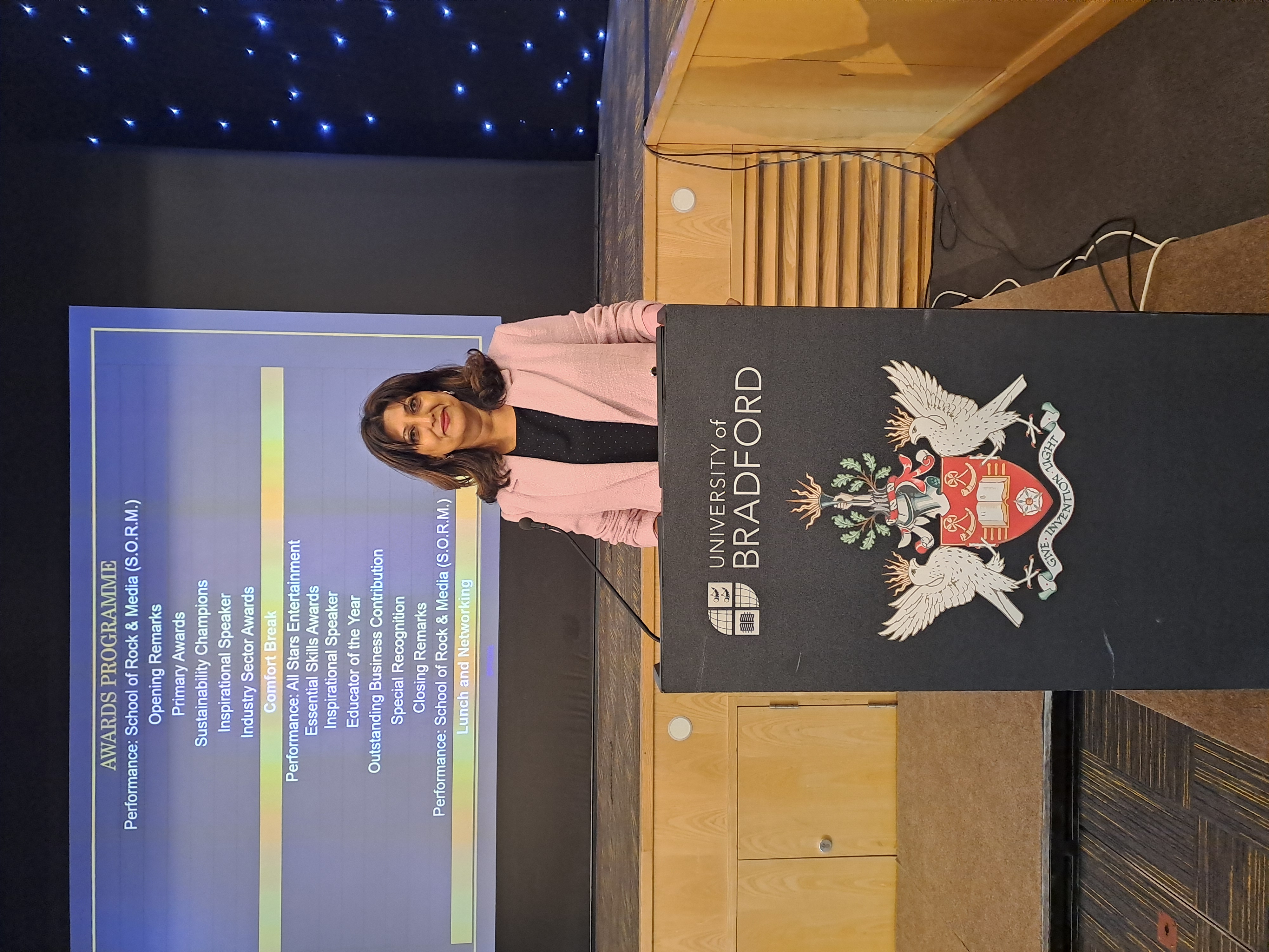 A woman stood at a University of Bradford lectern in front of a screen showing a programme of an event
