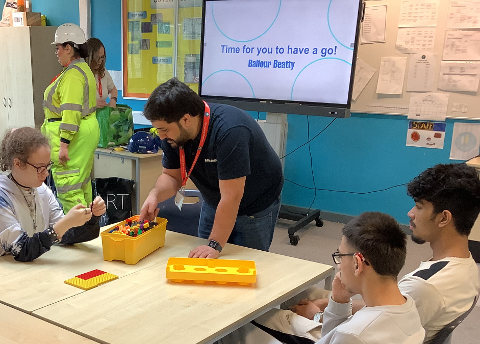 Young people sat at a classroom desk talking to a man wearing a lanyard