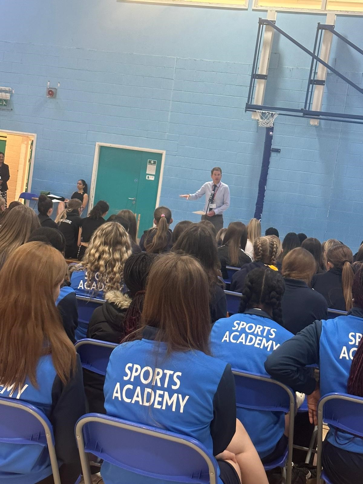 A large group of girls sat listening to a talk in a sports hall