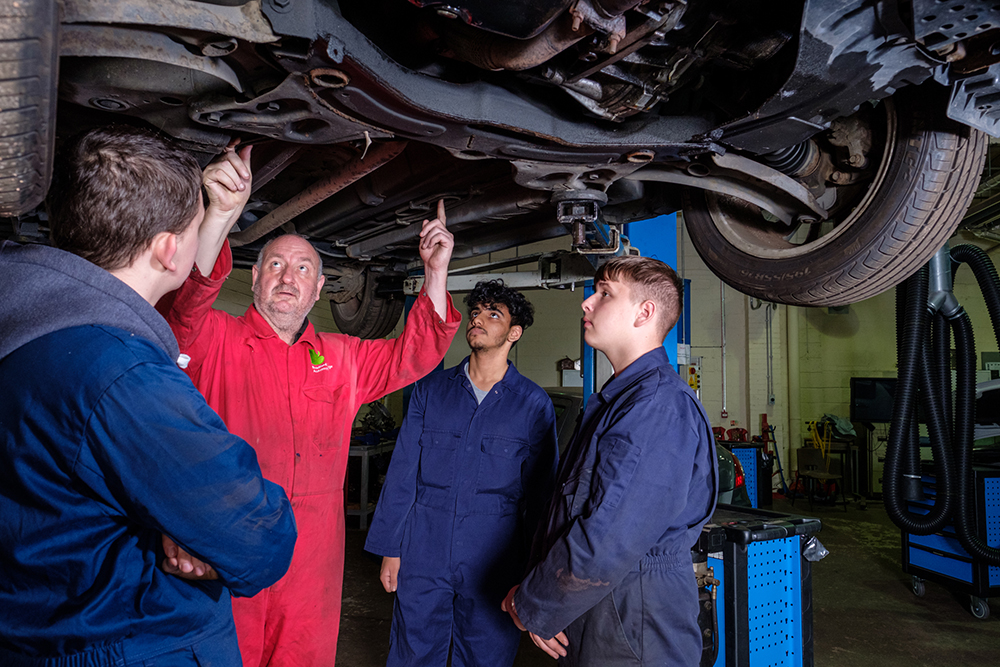 Three young men in overalls underneath a car, with an older man pointing out bits of the car exhaust