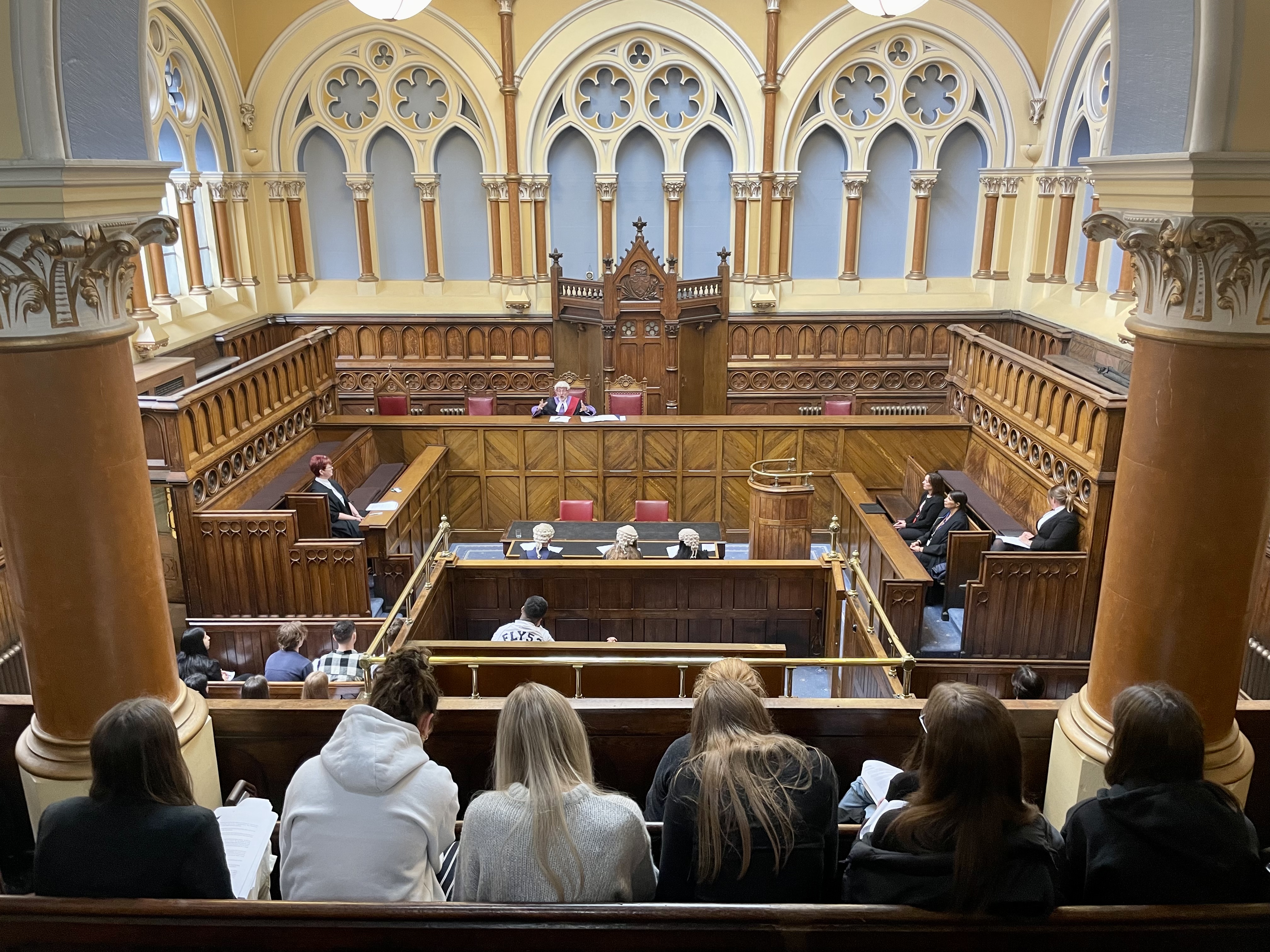 A judge sat at the front of a courtroom, facing two people in barristers' wigs and gowns, with a group of young people watching
