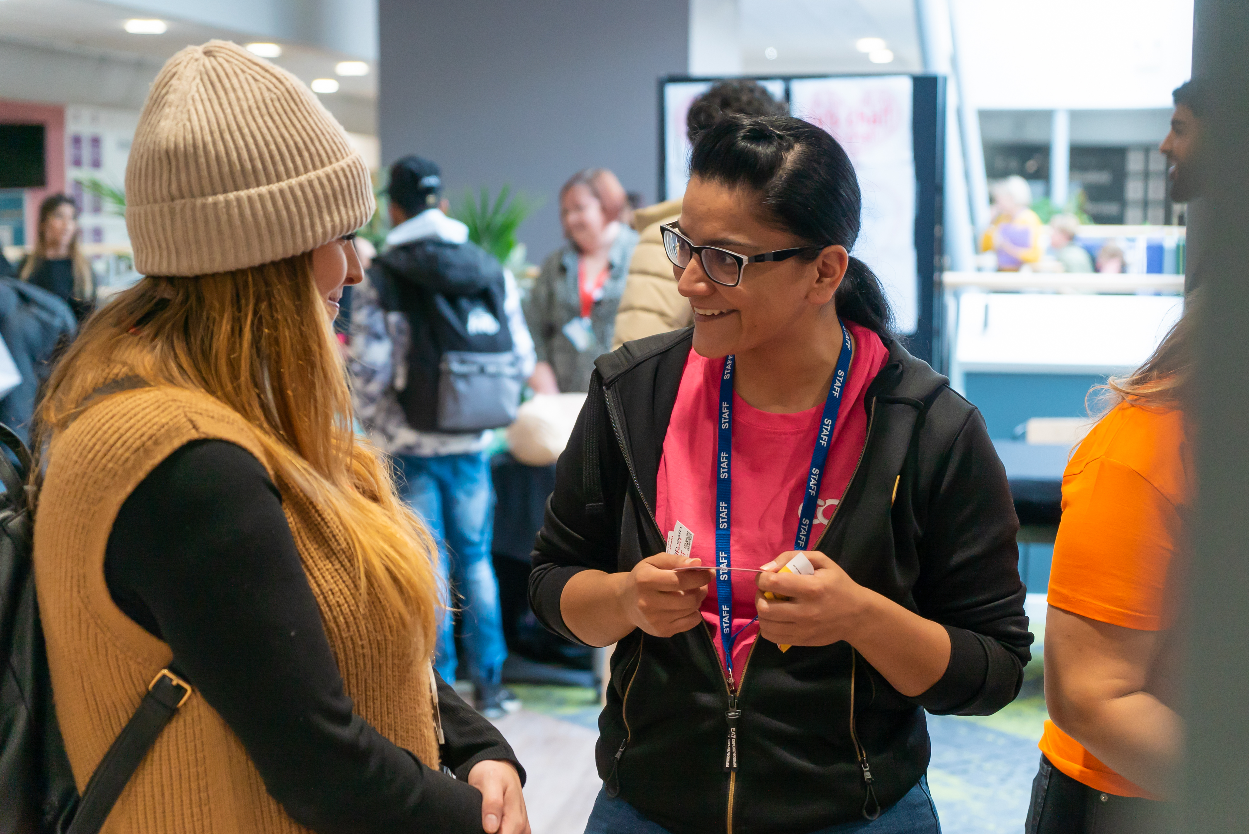 Woman in ID badge talking to woman. In background, more people talking
