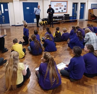 A group of young children sat on the floor in a hall listening to a talk from a man with a guide dog
