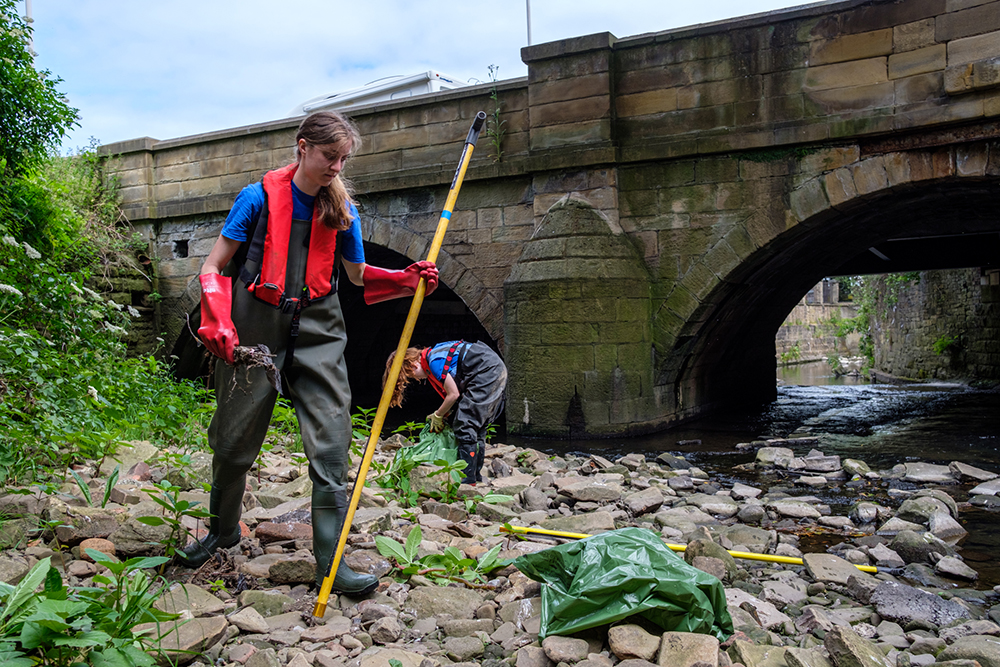 Two people in protective clothing collecting rubbish on a river bank