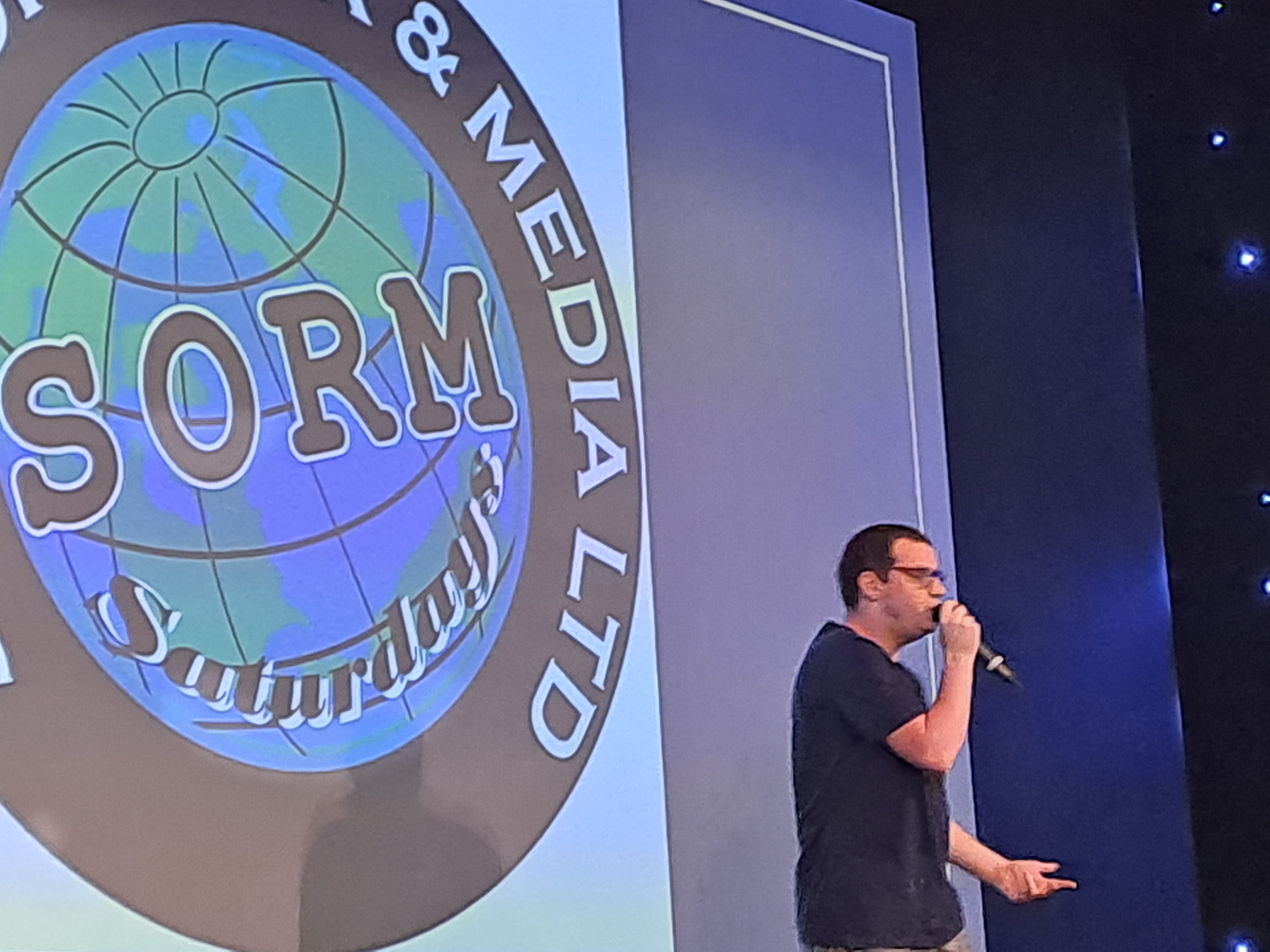A young man with microphone on stage in front of a sign saying School of Rock and Media