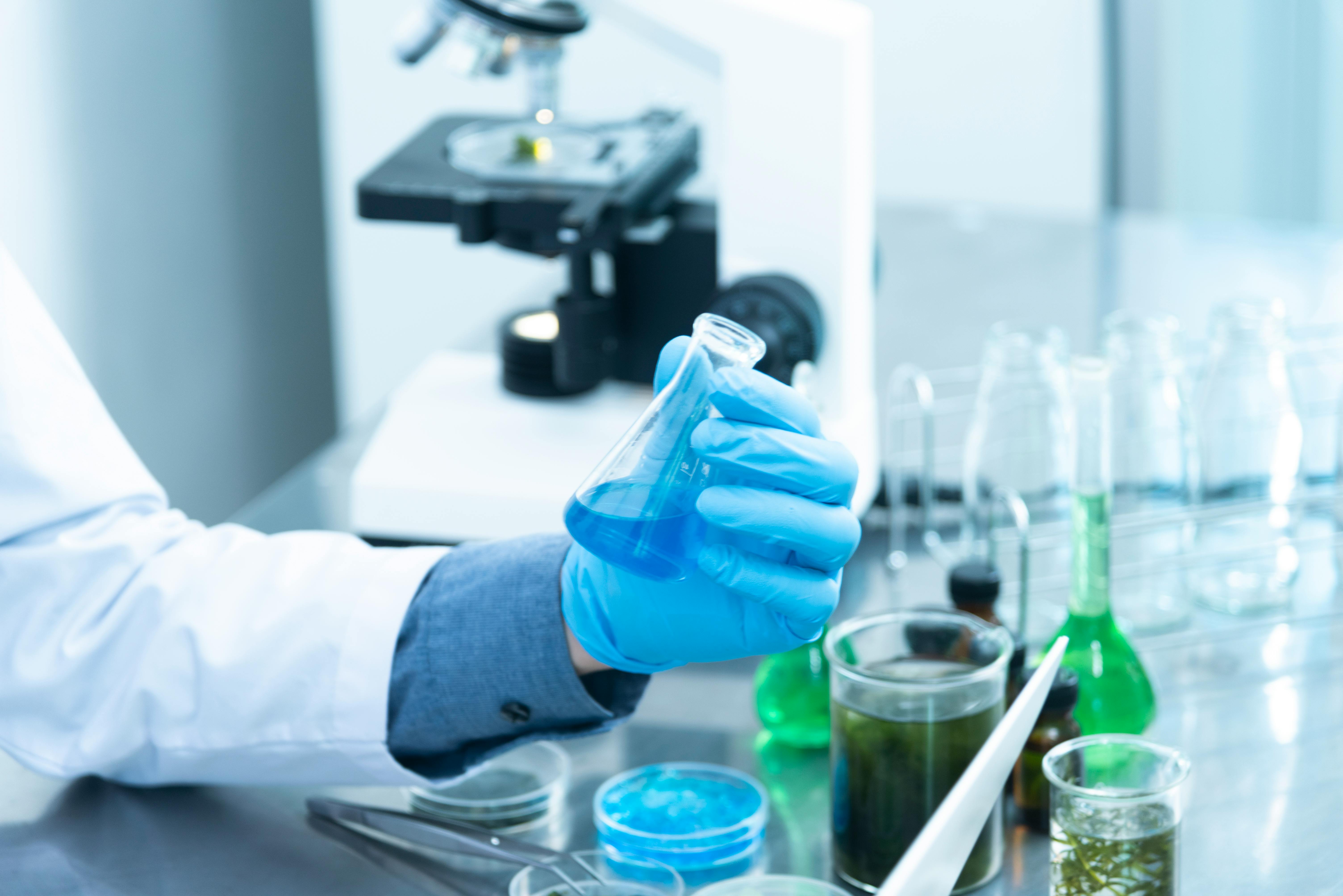 A gloved hand holding a chemical flask, with a microscope in the background