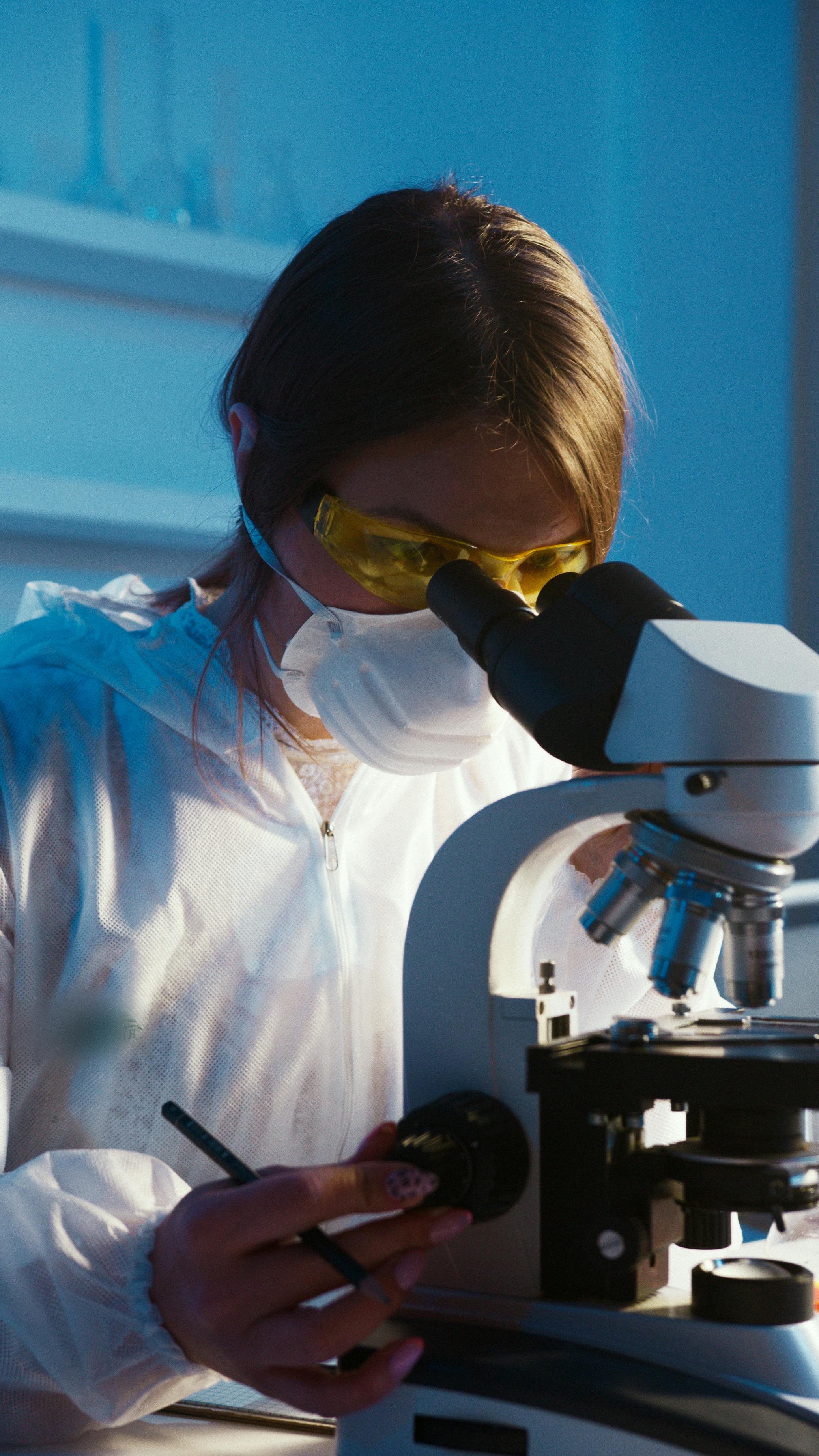 A woman in protective clothing examining something with a microscope