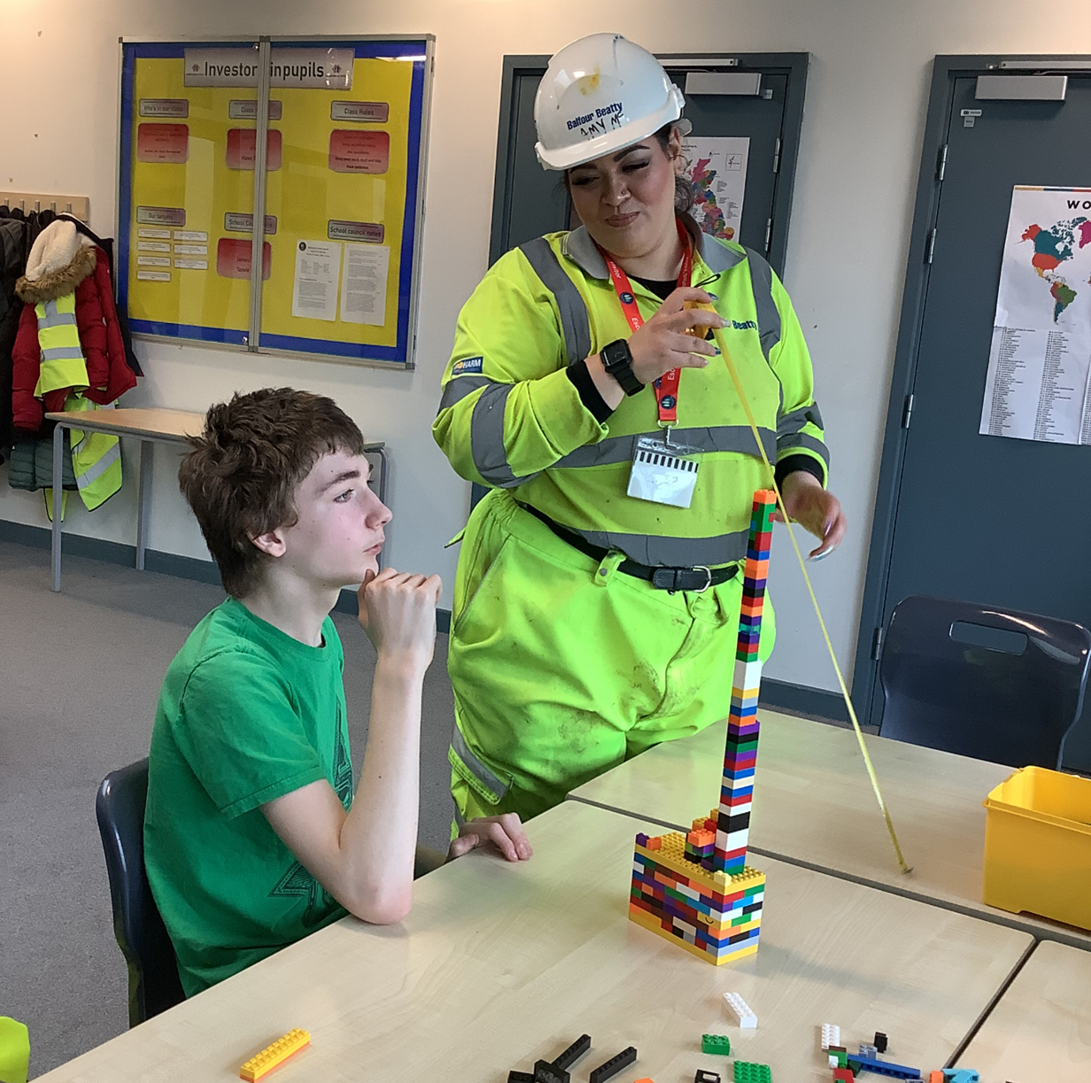 A woman wearing high vis and a hard hat talking to a young person building a tower of blocks
