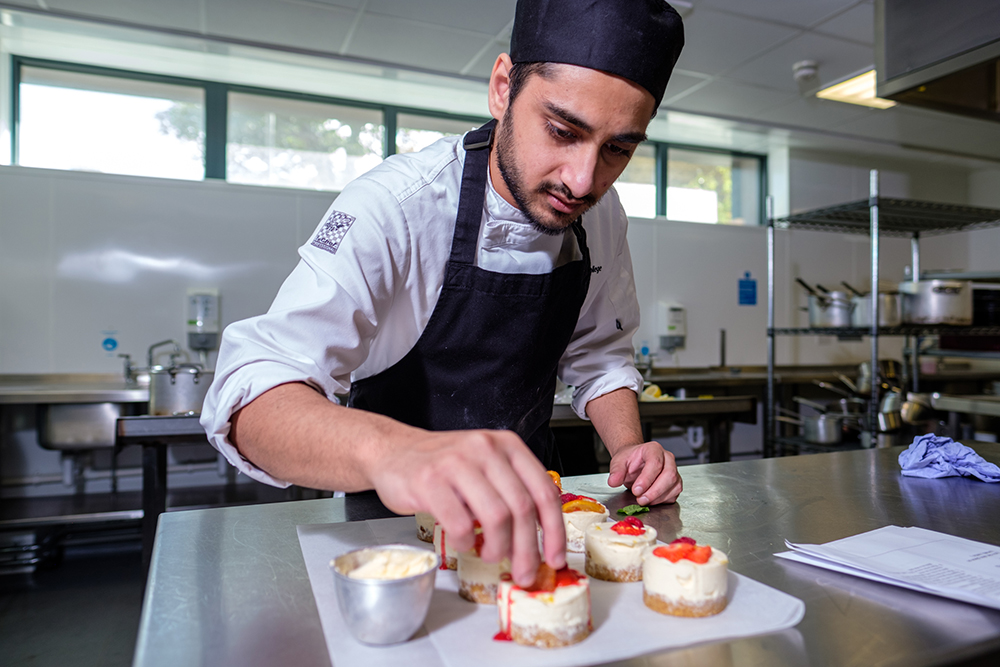 A young man wearing a chef's uniform putting fruit on top of a dessert