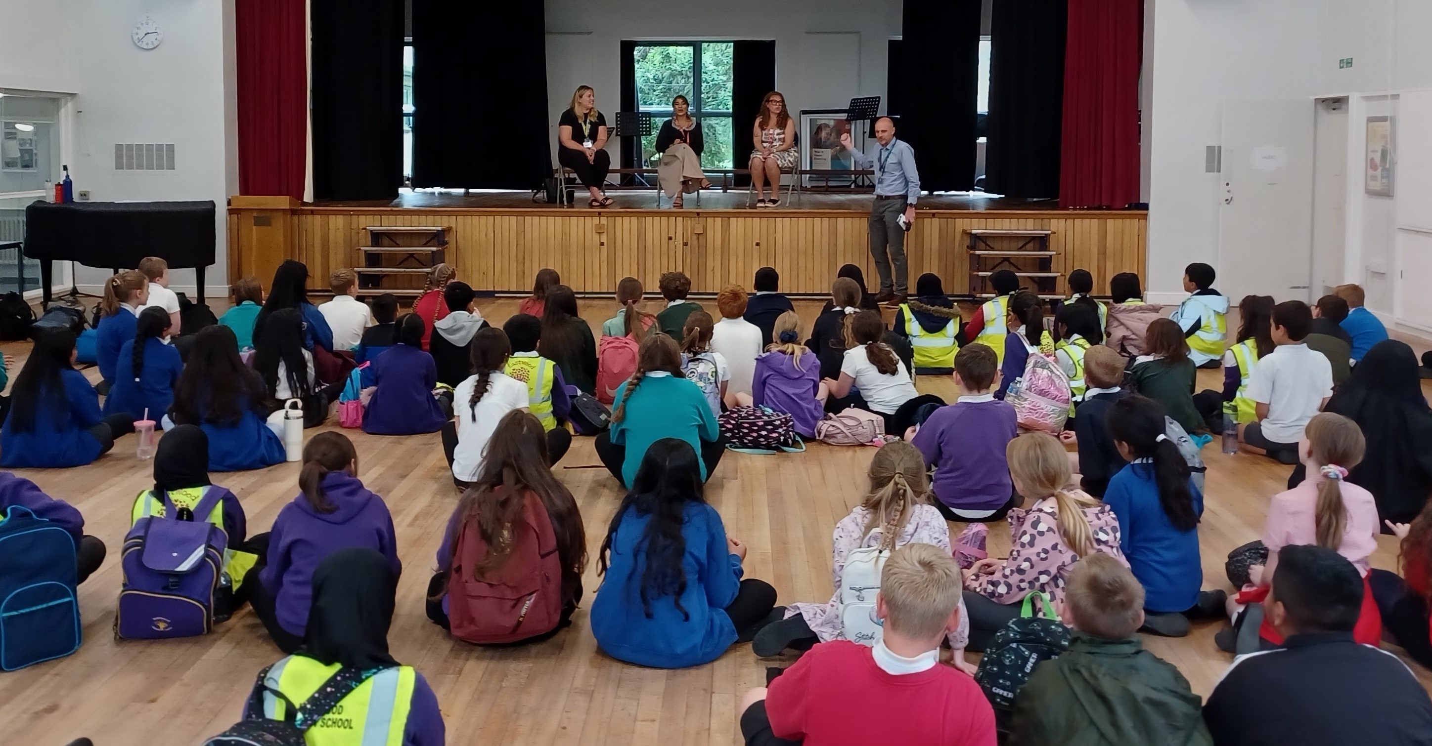 A large group of school children sat in a hall listening to three speakers